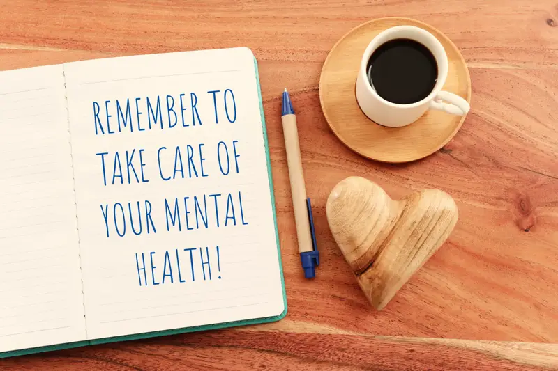 Notebook with a reminder to take care of your mental health, next to a coffee cup and heart-shaped ornament, promoting addiction treatment and mental health awareness.