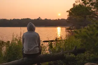 A person sitting alone at a lake during sunset, reflecting on their recent drug relapse.