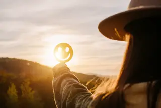 A woman holding a smiley face against the sunrise symbolizes her positivity and well-being.