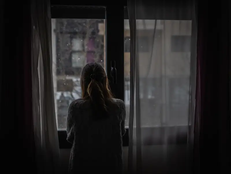 A woman stands by a window, looking out into a dreary, overcast scene, symbolizing the isolation and struggle often experienced during benzo withdrawal and addiction recovery.