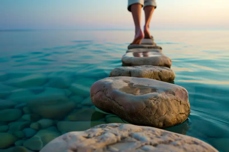 A close-up of a person walking on stepping stones over water, symbolizing the careful and deliberate steps taken in the journey to sobriety and recovery through AA.